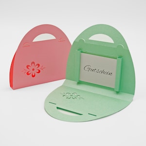 Könnte beinhalten: Zwei Geschenkkartenhalter aus Papier in Form einer Handtasche. Einer ist rosa mit einem roten Blumenmuster, der andere ist offen, grün und zeigt das Wort "Gutschein" auf einer weißen Karte. Beide haben einen Griff und dekorative Ausschnitte.