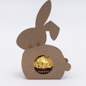 May include: A brown cardboard Easter bunny-shaped candy holder with a circular window. The window displays a gold-wrapped chocolate sweet. The bunny has long ears and a small tail.