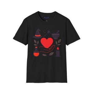 May include: Black t-shirt featuring a red heart held in a hand, surrounded by witch-themed illustrations. Includes a witch hat, magic wand, mortar and pestle, potion bottle, candle, and leaves. The design is in shades of red, purple, and brown.