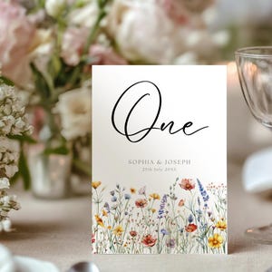 May include: A wedding table number card displaying the word "One" in black script. The card's base has a watercolour floral design with colourful wildflowers. The names "Sophia & Joseph" and the date are printed below.