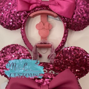 May include: Pink sequined Minnie Mouse ears headband with a large pink satin bow. The headband is displayed on a pink stand with a smaller pink Minnie Mouse head.
