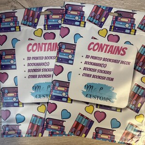 May include: Three white zippered pouches with a colorful book stack pattern. The pouches have a pink and white label that says "CONTAINS" and lists the items included: 3D printed bookshelf decor, bookmark(s), bookish stickers, and other bookish items. The label also has the text "M&L CUSTOMS" in a blue and white brushstroke font.