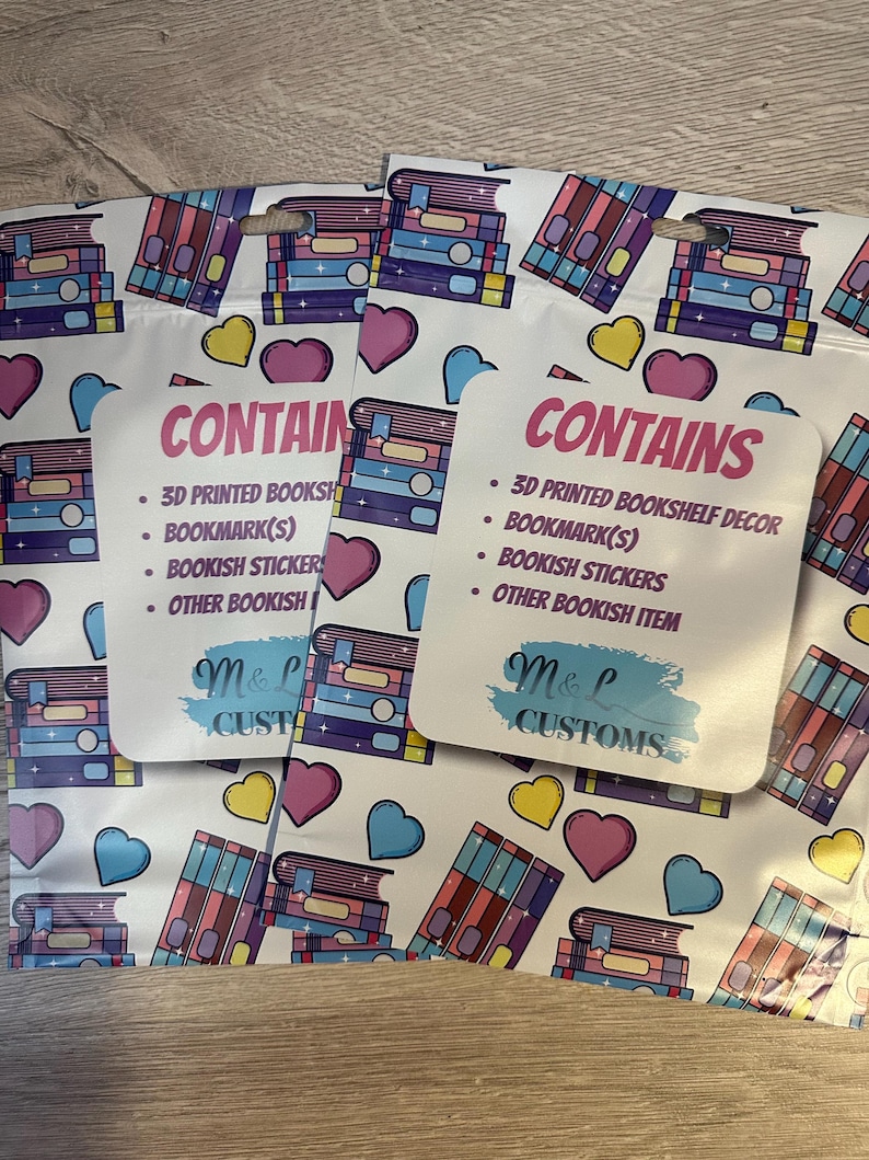 May include: Three white zippered pouches with a colorful book stack pattern. The pouches have a pink and white label that says "CONTAINS" and lists the items included: 3D printed bookshelf decor, bookmark(s), bookish stickers, and other bookish items. The label also has the text "M&L CUSTOMS" in a blue and white brushstroke font.