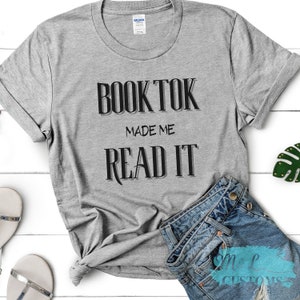 Puede incluir: Camiseta gris con el texto "BookTok Made Me Read It" en letras negras.