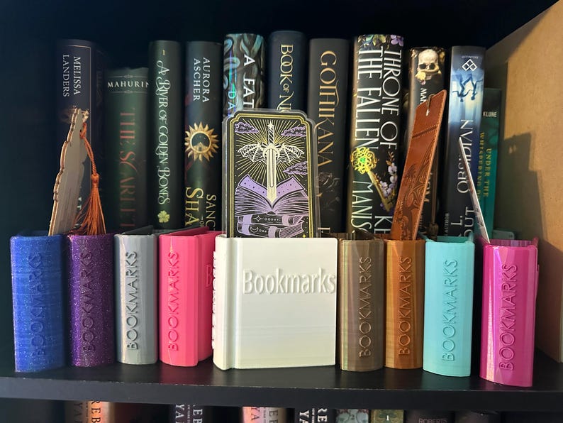 K&ouml;nnte beinhalten: Ein B&uuml;cherregal mit einer Auswahl an B&uuml;chern, darunter "The Fallen Manis" und "Gohikana". Auf dem Regal befinden sich mehrere farbige Lesezeichen, darunter ein wei&szlig;es mit der Aufschrift "Bookmarks".