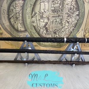 May include: Three wizarding wands displayed on a triangular stand. The wands are black, brown, and gray. The stand is made of a 3D printed material. The wands are displayed in front of a map of Diagon Alley from the Harry Potter series.
