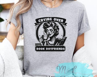 May include: Grey t-shirt with a black and white illustration of a woman crying over a book. The text "Crying Over Book Boyfriends" is printed on the shirt.