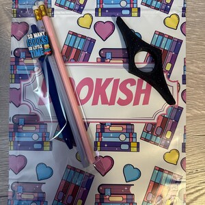 May include: A white zippered pouch with a bookish theme. The pouch has a pink and white background with a repeating pattern of stacks of books, hearts, and stars. The pouch has a black bookmark and three pencils inside. The text "SO MANY BOOKS SO LITTLE TIME" is printed on a small tag.