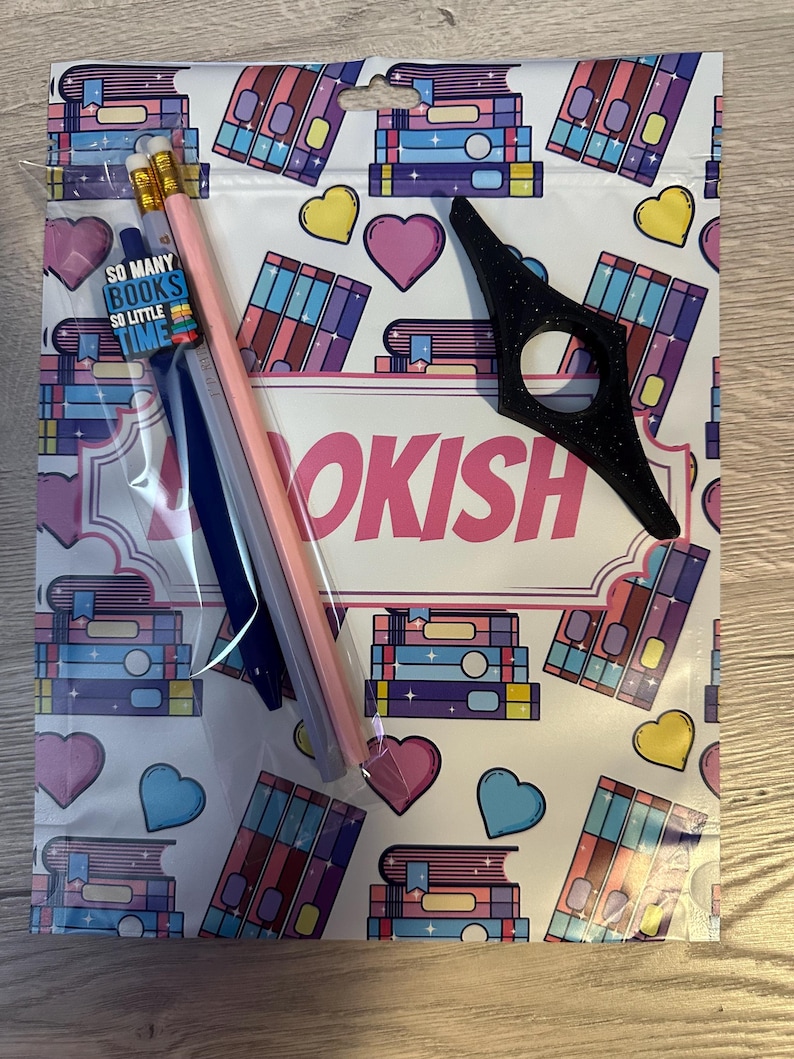 May include: A white zippered pouch with a bookish theme. The pouch has a pink and white background with a repeating pattern of stacks of books, hearts, and stars. The pouch has a black bookmark and three pencils inside. The text "SO MANY BOOKS SO LITTLE TIME" is printed on a small tag.