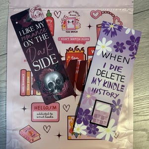 May include: Two purple and white bookmarks with funny sayings. The first bookmark says "I like my romance on the dark side" with a skull and crossbones graphic. The second bookmark says "When I die delete my kindle history".
