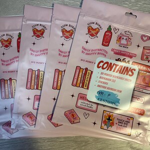 May include: Pink and white packaging with a variety of stickers and a bookmark. The stickers feature illustrations of books, hearts, and a sword. The bookmark is a pink and white rectangle with the text "CONTAINS" and a list of items included. The packaging also features the text "SLOW BURN?" and "EASILY DISTRACTED BY SMUTTY BOOKS".