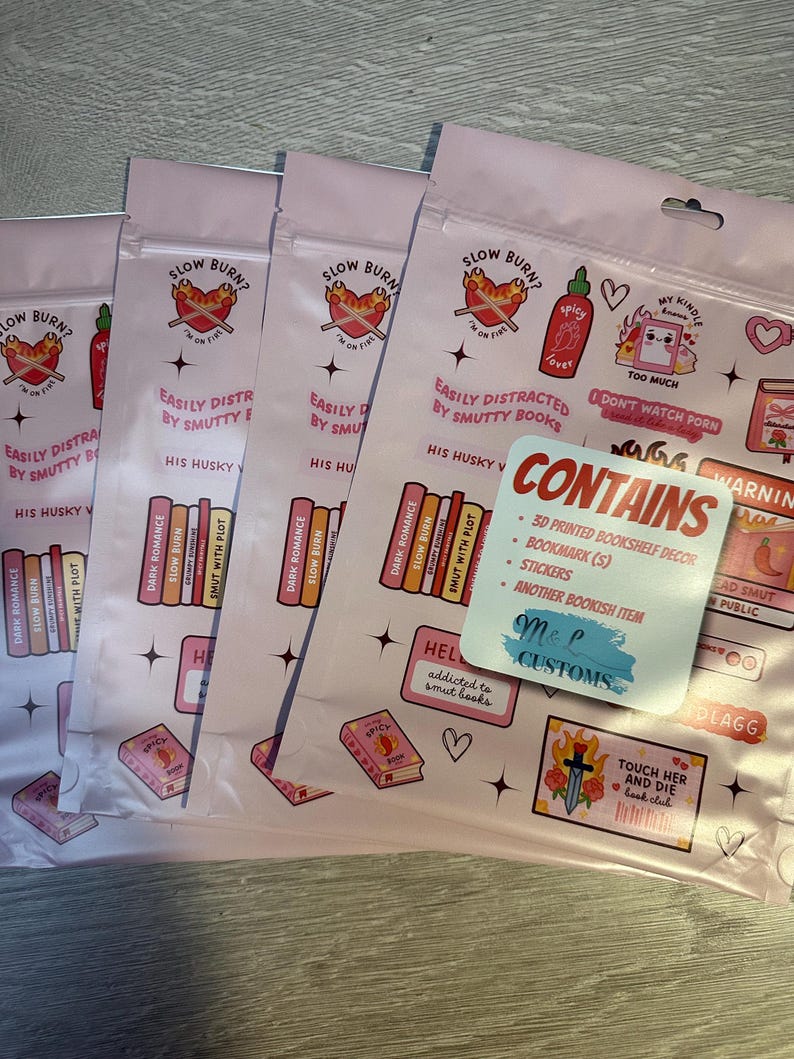 May include: Pink and white packaging with a variety of stickers and a bookmark. The stickers feature illustrations of books, hearts, and a sword. The bookmark is a pink and white rectangle with the text "CONTAINS" and a list of items included. The packaging also features the text "SLOW BURN?" and "EASILY DISTRACTED BY SMUTTY BOOKS".