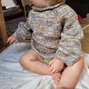 Op de afbeelding: Een gebreide trui met lange mouwen in een gemêleerd patroon van beige, bruin en lichtblauw garen. Het kledingstuk heeft een eenvoudige ronde halslijn en geribbelde boorden bij de polsen, gemaakt met een textuursteek voor een gespikkeld effect.