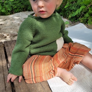 May include: A child in a green knitted jumper with a cream-coloured patch and orange striped trousers. The jumper has long sleeves and a crew neck. The trousers have vertical stripes in shades of orange, brown, and cream.