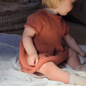 Könnte beinhalten: Ein gestricktes Kleid mit kurzen Ärmeln in einem warmen, rötlich-braunen Farbton. Das Kleid hat einen runden Ausschnitt und ein einfaches, strukturiertes Strickmuster. Das Kleidungsstück ist für ein Baby oder Kleinkind konzipiert.
