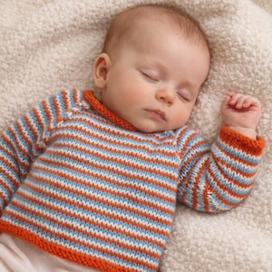 May include: A sleeping baby wearing a striped sweater. The sweater has horizontal stripes in orange, light blue, and white. The sweater has orange trim around the neck and cuffs. The baby is lying on a soft, cream-colored blanket.