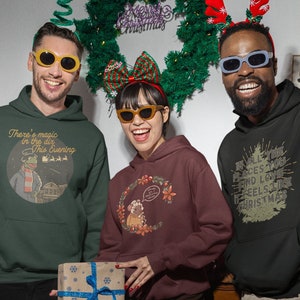May include: Three people wearing green, brown, and black hooded sweatshirts with Christmas-themed designs. The designs include a frog, a squirrel, and a Christmas tree. The people are wearing sunglasses and smiling.