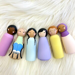 May include: Six painted wooden peg dolls with diverse skin tones, hairstyles, and outfits, including a doll in a wheelchair. The dolls are arranged on a white, fluffy surface. The dolls are designed with painted faces and clothing.