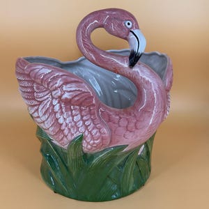 May include: A ceramic planter shaped like a pink flamingo with a curved neck and head. The flamingo is depicted standing in green grass, with its wings folded against its body. The interior of the planter is a smooth, light gray, glossy finish.