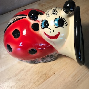 May include: A ceramic ladybug-shaped planter with a smiling face, blue eyes, and black antennae. The ladybug's shell is red with black spots, and the face is cream-colored. The planter has a black rim and is likely vintage.