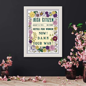 May include: A framed poster with the text "IRISH CITIZEN" and "VOTES FOR WOMEN NOW! DAMN YOUR WAR!" surrounded by floral illustrations. The poster is in a white frame, with pink flowers in vases and scattered on a wooden surface.