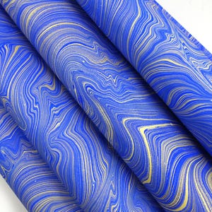 May include: Rolled sheets of paper with a marbled design. The paper has a vibrant blue background with swirling gold and cream-coloured veins, creating a luxurious and artistic appearance. The paper is likely used for crafts or decorative purposes.