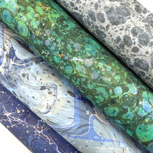 May include: A close-up of several rolls of marbled paper in various colors. The rolls display a range of patterns, including swirls and spots, in shades of blue, green, gray, and white. The paper appears to be high-quality and suitable for crafts.