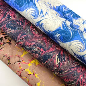 May include: Close-up of four rolls of marbled paper in various colors. The rolls display intricate patterns in shades of blue, white, pink, black, yellow, and brown. The paper has a swirling, organic design, suitable for crafts or decorative purposes.