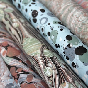 May include: Several rolls of paper featuring intricate marbled patterns in various colour palettes. The designs include swirling black, sage green, and terracotta lines, alongside a light blue sheet with scattered black and grey circular spots.