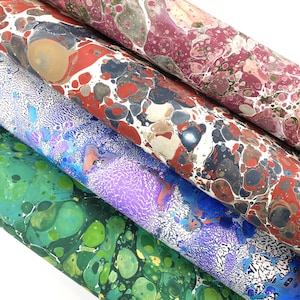 May include: Close-up of several rolls of marbled paper in various colors. The rolls display a mix of colors including green, blue, red, and purple, with swirling patterns. The paper appears to be high-quality, suitable for crafts or artistic projects.