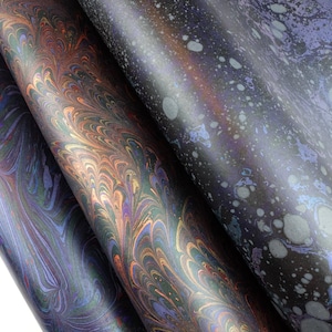 May include: Three rolls of paper featuring distinct marbled patterns. The left and center rolls display intricate, swirling metallic copper, blue, and purple designs. The right roll features a dark, speckled pattern with light gray circular spots.