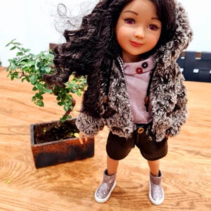 May include: A doll with dark curly hair, wearing a faux fur hooded jacket, a pink shirt, black shorts, and silver glitter boots. A small bonsai tree in a wooden pot is to the left of the doll.