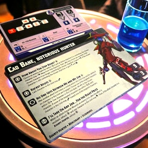 Peut inclure: Une scène de table avec une carte de jeu avec le texte "CAD BANE, NOTORIOUS HUNTER" et une illustration de personnage. Une boisson bleue dans un verre et une autre carte de jeu sont également sur la table. Les cartes sont pour un jeu de société.