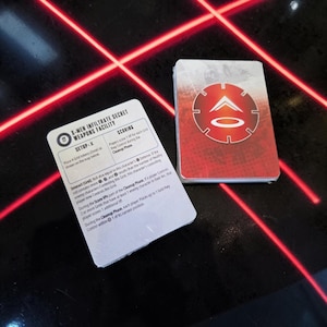 May include: Two stacks of game cards. One card displays the text "X-MEN INFILTRATE SECRET WEAPONS FACILITY" and game instructions. The other card has a red and white circular design. The cards are on a black surface with red laser-like lines.