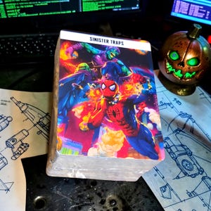 May include: A rectangular deck of playing cards featuring a vibrant illustration of Spider-Man and the Green Goblin with the text SINISTER TRAPS printed at the top. The artwork displays bright red, blue, purple, and green colours with explosive effects.
