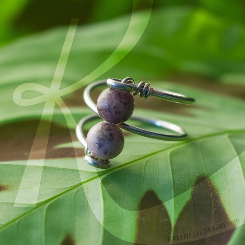 Op de afbeelding: Een zilveren draadring met twee kleine, gevlekte, lichtpaarse en grijze kralen. De ring ligt op een groen blad.
