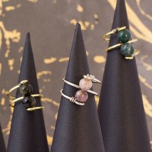 Op de afbeelding: Drie zilveren ringen met verschillende gekleurde stenen op zwarte kegelvormige displays. De ringen hebben elk twee stenen. De stenen zijn groen, roze en zwart.