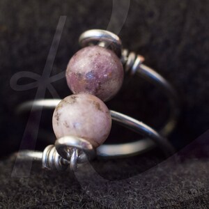 Op de afbeelding: Een zilveren ring met twee roze en grijze edelsteen kralen. De ring is om zichzelf gewikkeld en heeft een gedraaid draadontwerp.