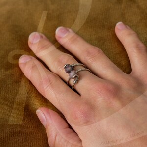 Op de afbeelding: Een zilveren ring met twee paarse en grijze edelsteen kralen. De ring zit aan een vinger.