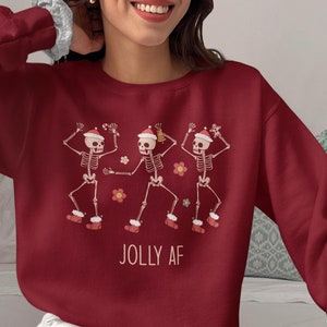 May include: Burgundy crewneck sweatshirt featuring a festive design of three dancing skeletons wearing Santa hats and red boots. The phrase "JOLLY AF" is printed below the skeletons. The sweatshirt is a comfortable, casual style.