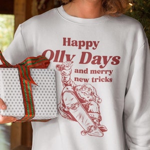 May include: White sweatshirt with the text "Happy Olly Days and merry new tricks" and a graphic of Santa Claus skateboarding. The person is wearing a Santa hat and holding a wrapped gift with a red and green ribbon.