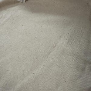 May include: A close-up of a beige t-shirt with a small, white tag that reads "RUSSELL" and "180M S". The t-shirt has a few small, dark spots.