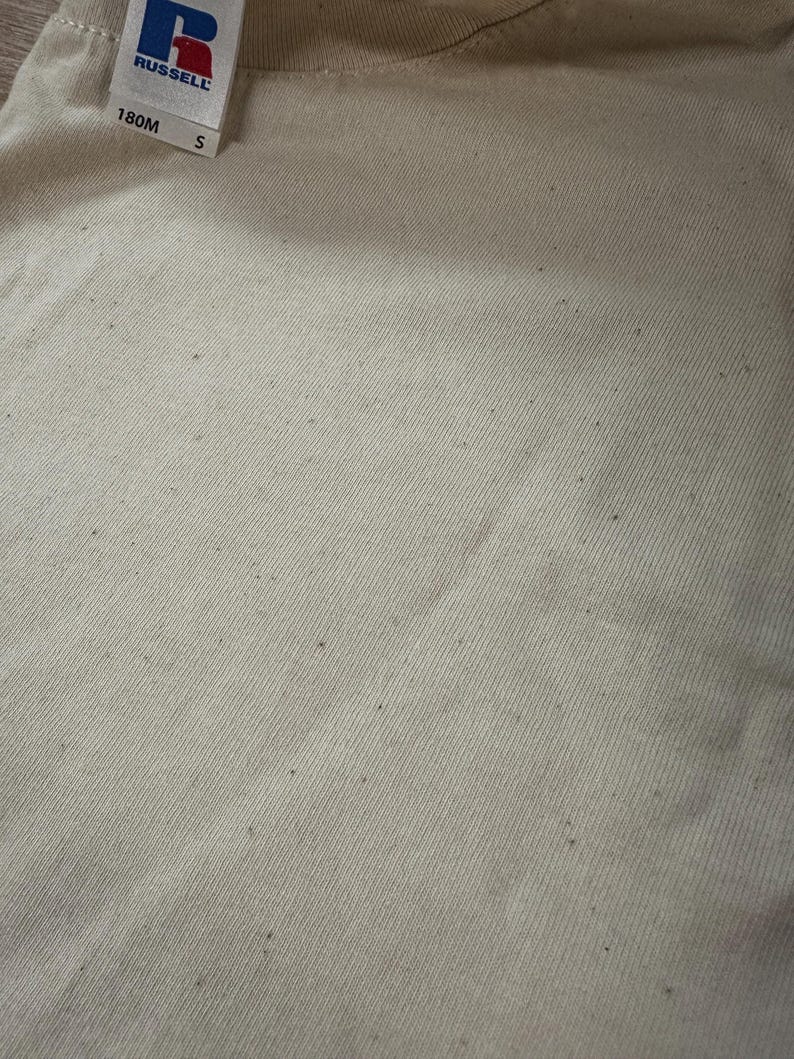 May include: A close-up of a beige t-shirt with a small, white tag that reads "RUSSELL" and "180M S". The t-shirt has a few small, dark spots.