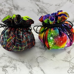 May include: Two drawstring bags for dice, one with a black and neon colored plaid pattern and the other with a rainbow galaxy print.
