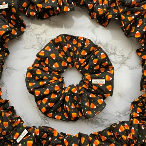 May include: Black scrunchies with a candy corn pattern. The fabric features orange, yellow, and white candy corn shapes on a black background. The scrunchies are gathered and voluminous, with a small white tag that reads "Molly Twilight".