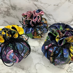 May include: Three drawstring bags with a stained glass pattern featuring a castle, a rose, and a clock. The bags are made of fabric and have a black drawstring. The bags are lined with different colors of fabric: yellow, pink, and blue.