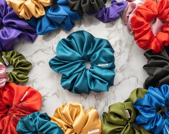 May include: A collection of colorful satin scrunchies in various colors, including teal, red, purple, green, black, and gold. The scrunchies are arranged in a circle on a white marble surface.