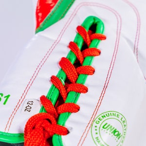 May include: Close-up of a white boxing glove with red and green accents. The glove is laced with red and green laces. The glove has a label that says "12-OZ" and a stamp that says "GENUINE LEATHER UNION HAND MADE".