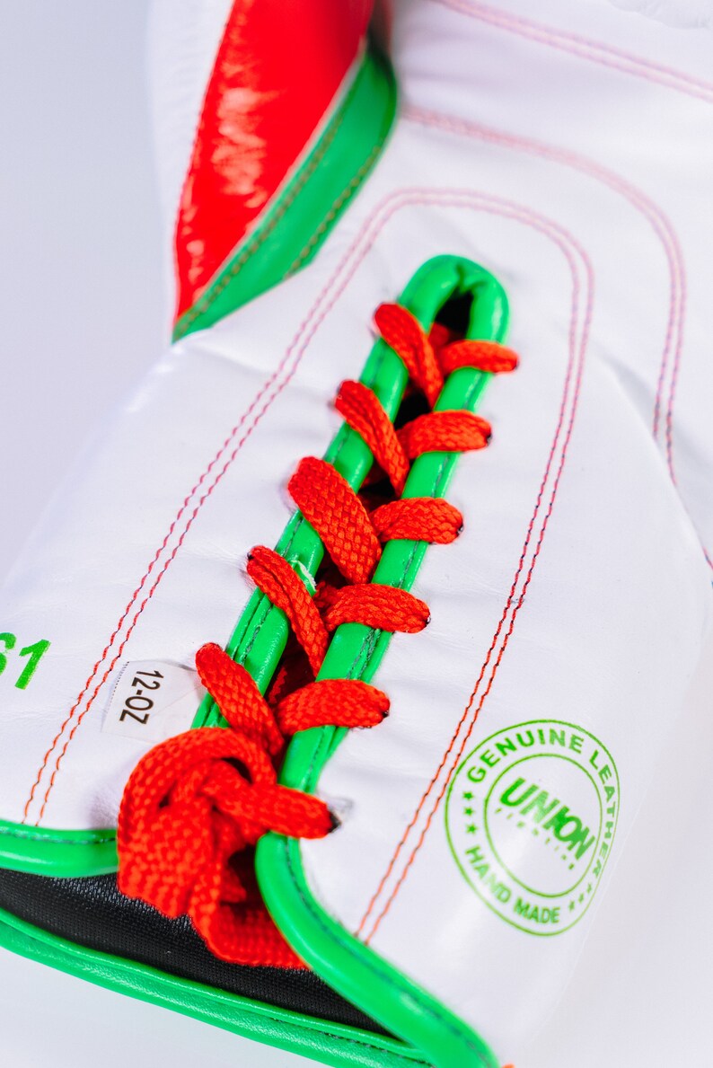 May include: Close-up of a white boxing glove with red and green accents. The glove is laced with red and green laces. The glove has a label that says "12-OZ" and a stamp that says "GENUINE LEATHER UNION HAND MADE".