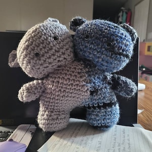 May include: Two crocheted stuffed animals, one gray and one blue, are standing together. The gray animal is in front of the blue animal. The gray animal has a simple design, while the blue animal has a more intricate design with black accents.